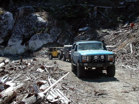Blue 1980s Ford Bronco with brush guard and snorkel on a debris-strewn dirt road.