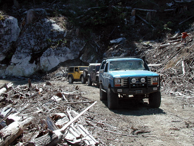 Blue 1980s Ford Bronco with brush guard and snorkel on a debris-strewn dirt road.