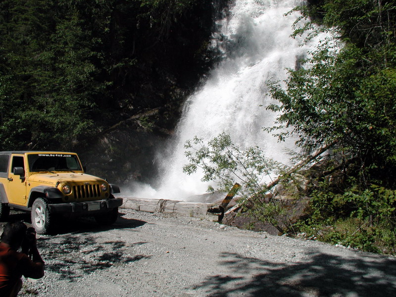 Yellow Jeep Wrangler Rubicon parked on gravel road by a waterfall.