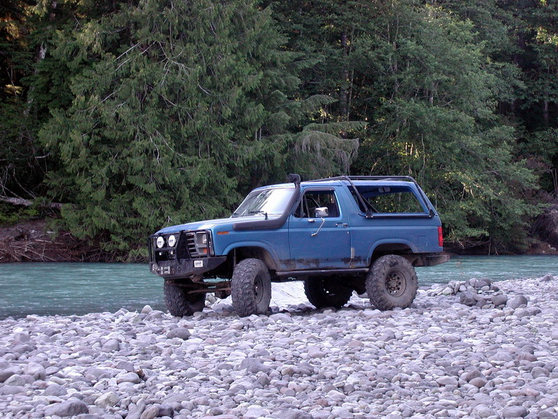 Blue Ford Bronco parked on a rocky riverbed with a forest and river behind it.