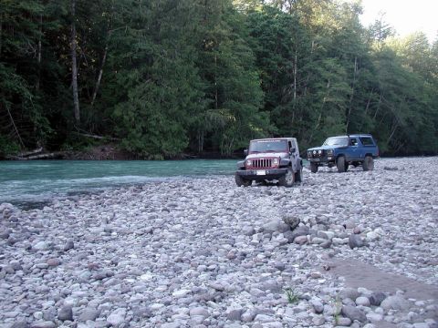 Maroon Jeep Wrangler and blue Ford Bronco on a rocky riverbank with a turquoise river and forest.