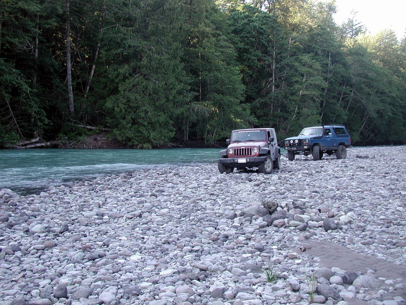 Maroon Jeep Wrangler and blue Ford Bronco on a rocky riverbank with a turquoise river and forest.