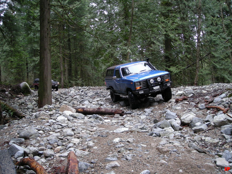 Blue 1980s Ford Bronco off-roading on a rocky trail in a forest.