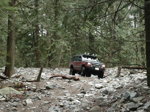 Red SUV driving on a rocky forest trail with roof-mounted lights