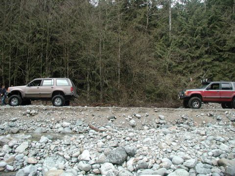 Two lifted SUVs parked on a rocky embankment with a forest behind them.