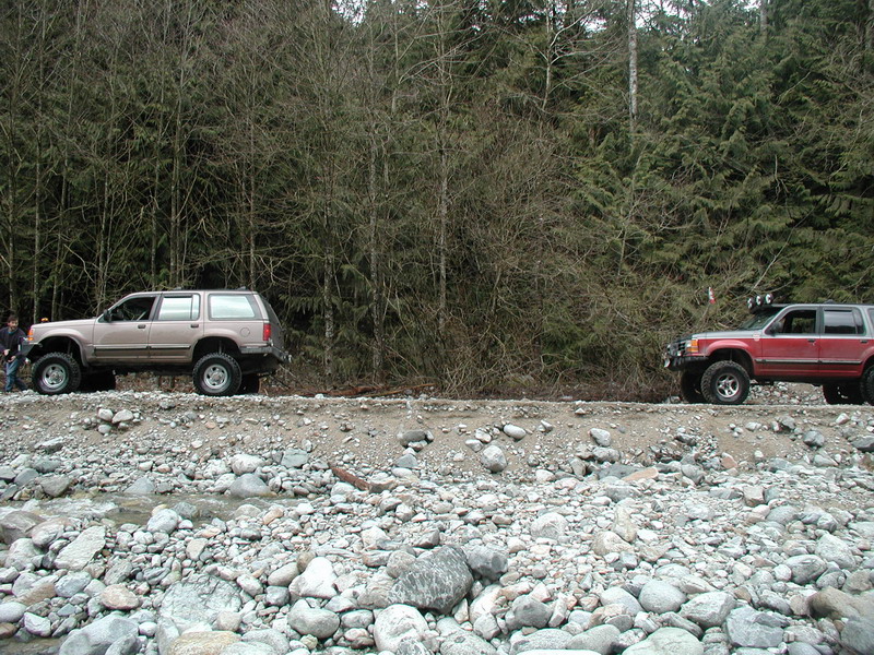 Two lifted SUVs parked on a rocky embankment with a forest behind them.