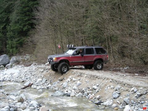Lifted red and gray SUV on a dirt path next to a rocky stream with auxiliary lights and a Canadian flag.