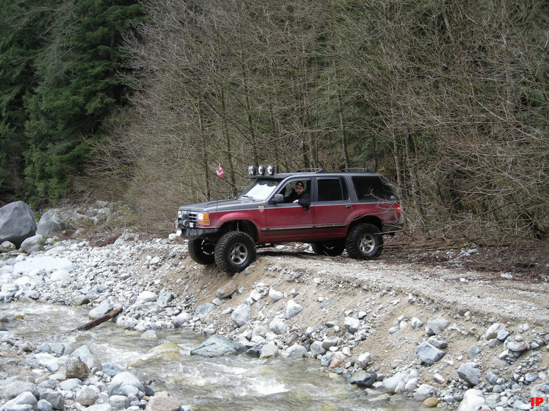 Lifted red and gray SUV on a dirt path next to a rocky stream with auxiliary lights and a Canadian flag.