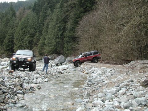 Two off-road SUVs in a rocky riverbed with a forest backdrop.