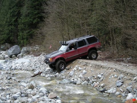 Red and gray SUV driving on a steep, rocky bank next to a stream.