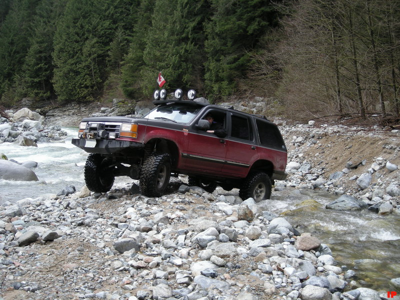 Lifted red SUV driving through a rocky river with a forest backdrop.