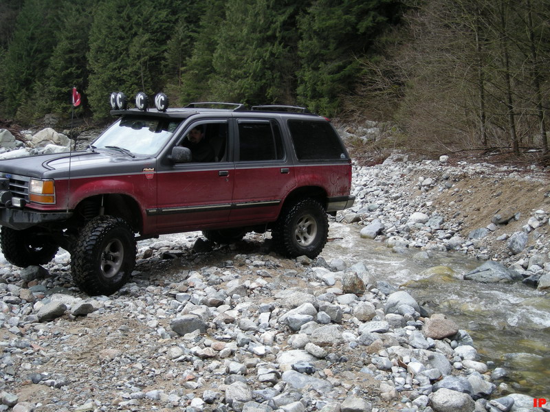 Lifted maroon and gray Ford Explorer driving through rocky stream with forest background.