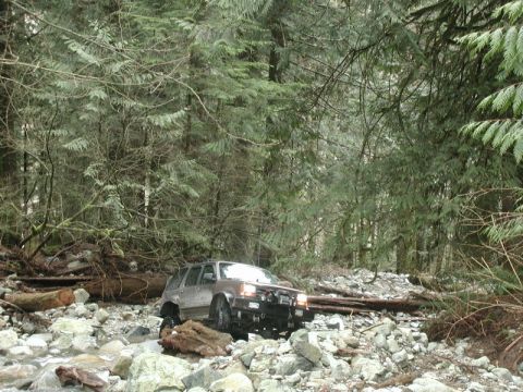 Brown SUV driving through a rocky stream in a forest.