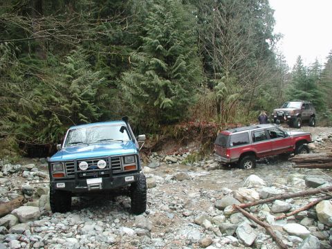 Blue Ford Bronco off-roading through a rocky stream in a forest.