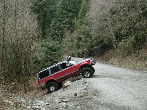 Red SUV tilted on a steep, loose gravel embankment next to a dirt road in a forest.
