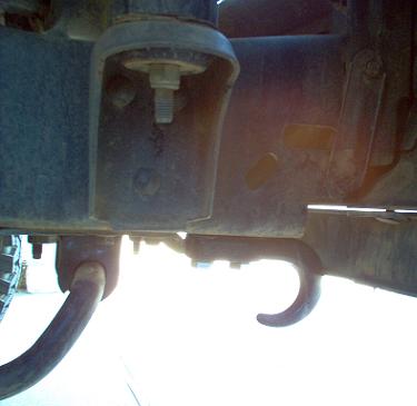 1988 Ford Bronco - Backlit low-angle shot under a truck's front end showing a bolted-on tow hook curving down off the frame rail, with a second hook or hose l…