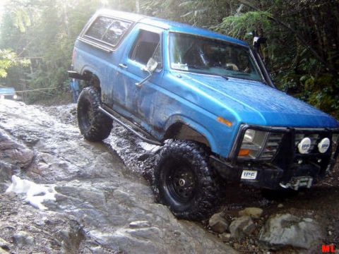 Blue 1980s Ford Bronco off-roading on a rocky, wet trail.