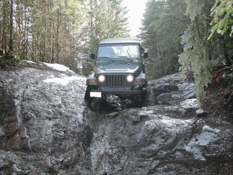 Dark green Jeep Wrangler climbing a steep rocky and muddy trail in a forest.