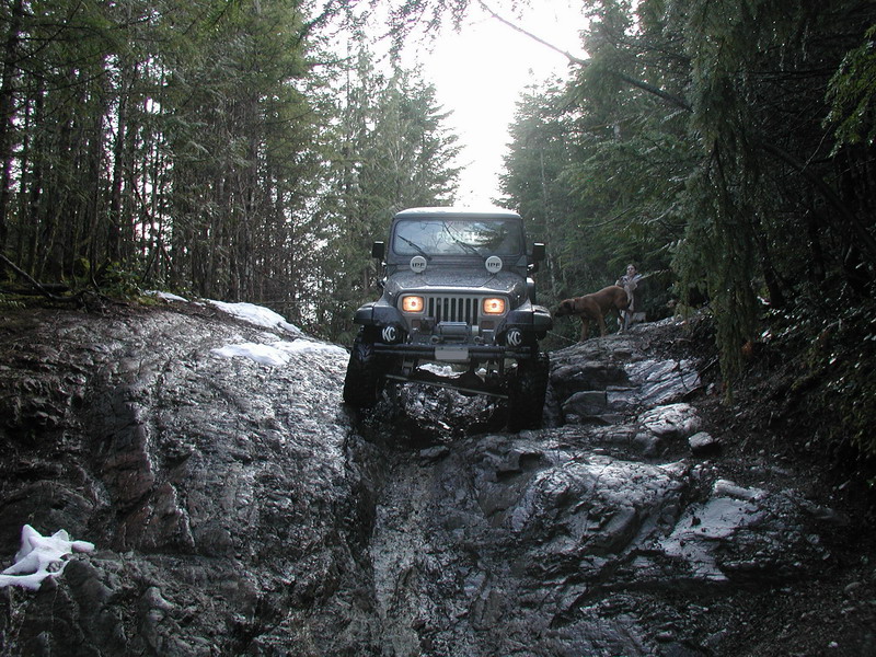 Jeep Wrangler driving on a rocky, snowy off-road trail in a forest.