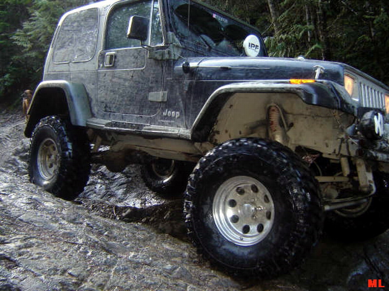 Mud-covered Jeep Wrangler with large tires on a rocky, wet trail.
