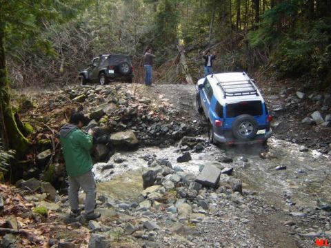 Blue and white SUV driving through a rocky stream in a forest.