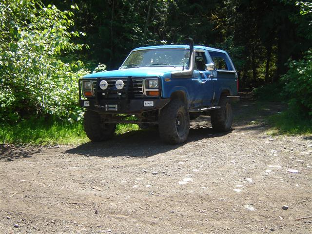 Blue 1985 Ford Bronco with snorkel and brush guard on a gravel road.