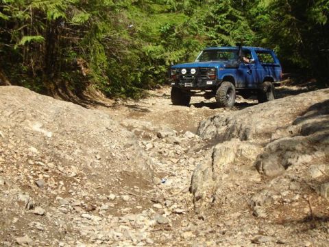 Blue 1980s Ford Bronco off-roading on a rocky trail.
