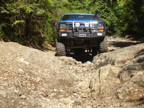 Blue Ford Bronco driving on a rocky off-road trail in a forest.