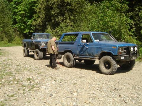 Two blue lifted Ford trucks, a Bronco and a pickup, parked on a gravel road with a man standing between them.