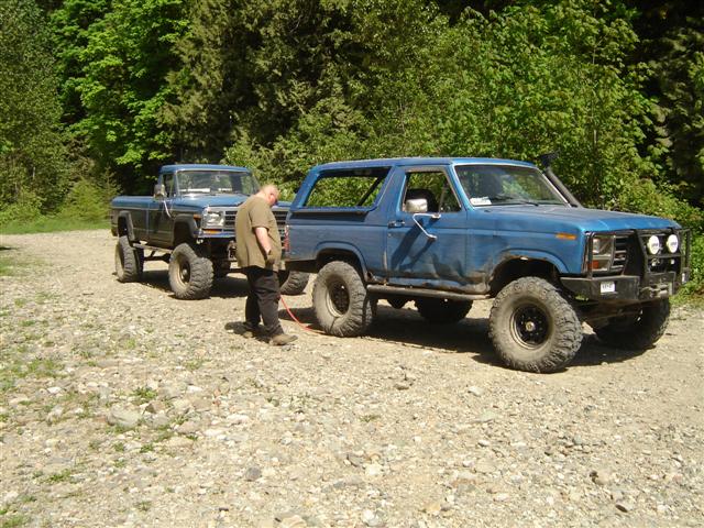 Two blue lifted Ford trucks, a Bronco and a pickup, parked on a gravel road with a man standing between them.