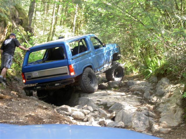 Blue Ford Bronco off-roading on a rocky trail in a forest.