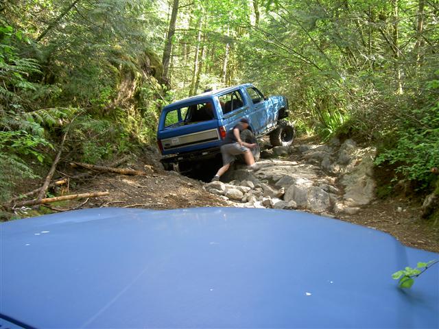 Blue Ford Bronco off-roading on a rocky, wooded trail.