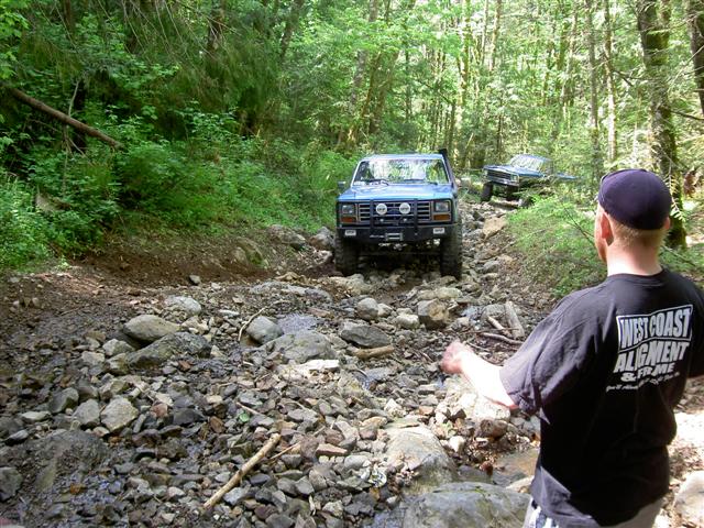 Blue Ford Bronco on a rocky trail in a forest, with a person in foreground.