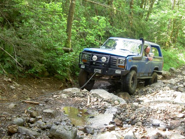 Blue 1980s Ford Bronco driving on a rocky trail with a snorkel and winch.