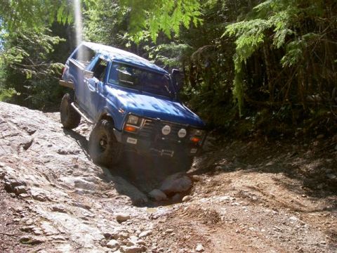 Blue 1980s Ford Bronco off-roading on a rocky, wooded trail.