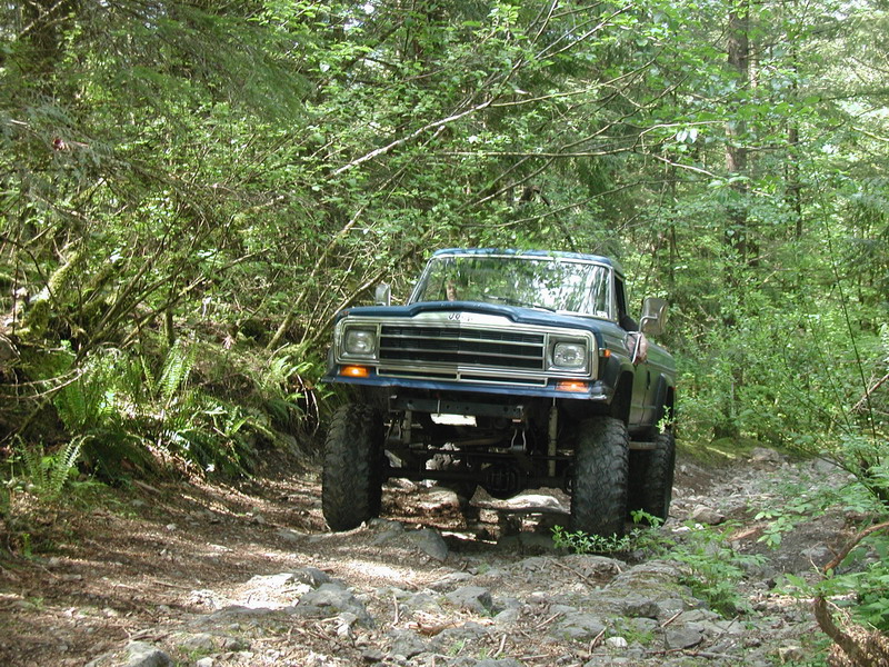 Blue Jeep pickup truck with large tires on a rocky forest trail.