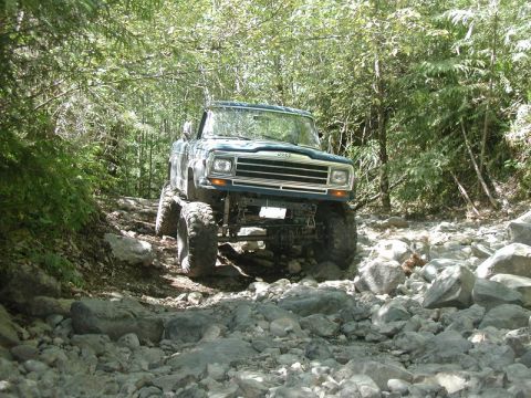 Dark blue Jeep pickup truck with large tires on a rocky, wooded trail.