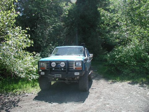 Lifted teal Ford Bronco with bull bar and snorkel parked on a gravel road in a forest.