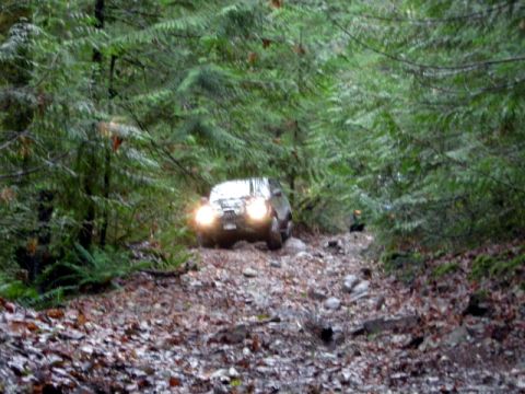 Dark SUV with headlights on driving on a rocky forest trail.