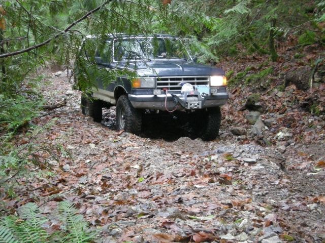 Dark blue Ford Bronco driving on a wooded trail with a winch on the front.