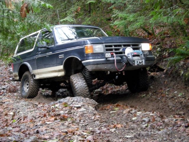 Dark blue and tan Ford Bronco off-roading on a muddy trail in a forest.