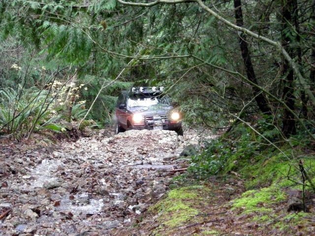 Dark SUV with roof lights driving on a rocky forest trail.