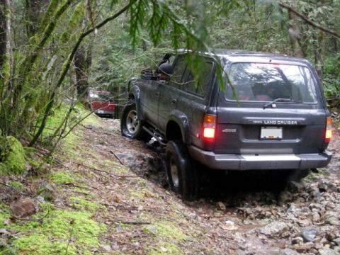 Dark gray Toyota Land Cruiser driving on a rocky, muddy forest trail.