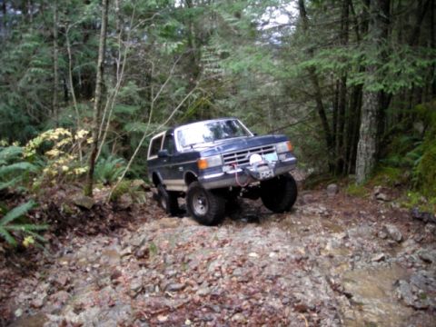 Dark blue Ford Bronco off-roading on a rocky, wooded trail.