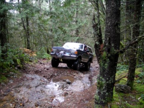 Dark Ford Bronco driving on a muddy, rocky forest trail.