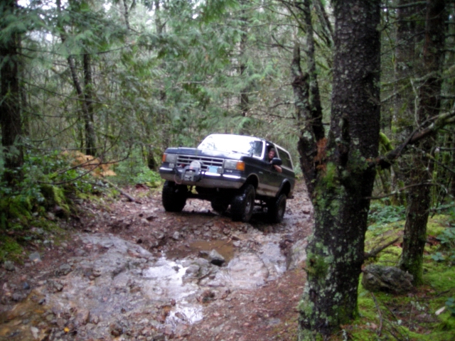 Dark Ford Bronco driving on a muddy, rocky forest trail.