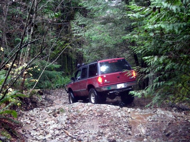 Red SUV driving on a muddy, rocky forest trail.