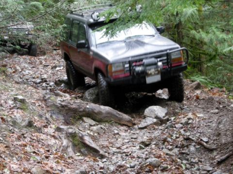 Dark red SUV with brush guard and winch driving on a rocky forest trail.