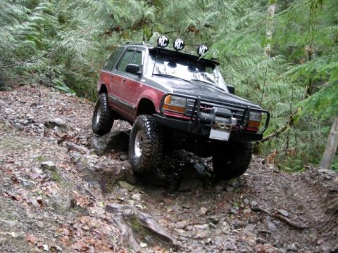 Red and gray off-road SUV with winch on a rocky, muddy forest trail.