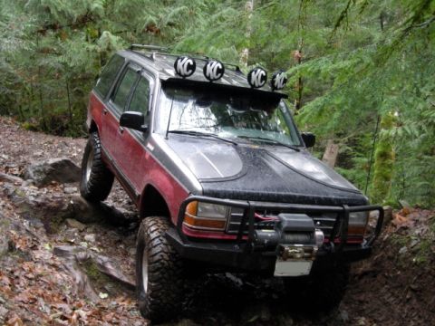 Red and black Ford Explorer off-roading on a rocky trail with winch and KC lights.